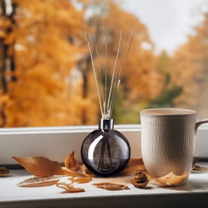 May include: A dark gray glass diffuser bottle with a silver cap and reeds, sitting on a white surface with dried leaves and a white ceramic mug.