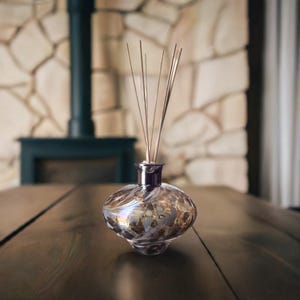 Könnte beinhalten: Ein dekorativer Glasdiffusor mit bauchigem Körper und dunkelviolettem Hals. Das Glas hat ein wirbelndes, irisierendes Muster mit goldenen und braunen Akzenten. Dünne, hellfarbene Stäbchen erstrecken sich nach oben vom Hals. Der Diffusor steht auf einer dunklen Holzoberfläche.