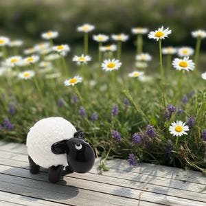 May include: A small glass figurine of a sheep with a textured white body and smooth black head, ears, and legs. The sheep has two white eyes with black pupils and is positioned on a light-coloured wooden surface with a blurred floral background.
