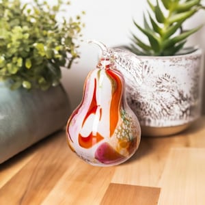 Può includere: Un ornamento a forma di pera in vetro con un design a spirale in tonalità di bianco, arancione, rosso e rosa. La pera ha uno stelo in vetro trasparente.