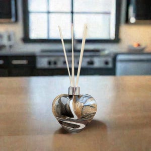 Op de afbeelding: Een hartvormige glazen diffuserfles met een zilveren dop en zwarte, witte en gouden marmering. De fles is gevuld met een heldere vloeistof en heeft houten diffuserstokjes die in de bovenkant zijn gestoken.