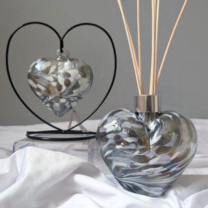 Op de afbeelding: Een hartvormige glazen diffuserfles met een zilverkleurige metalen bovenkant, gevuld met houten rietjes. Een bijpassend hartvormig glazen ornament hangt in een zwart metalen hartvormig frame. Het glas heeft een wervelend patroon van wit, grijs en helder.