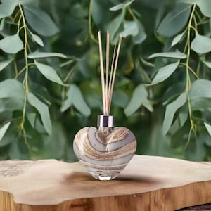 Könnte beinhalten: Eine herzförmige Glas-Diffusorflasche mit silbernem Hals und Holzstäbchen. Die Flasche hat ein grau-braunes Wirbelmuster. Der Hintergrund zeigt grüne Blätter, und der Diffusor steht auf einer Holzoberfläche.