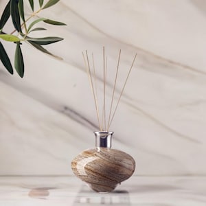 Op de afbeelding: Een ovale diffuserfles met een zilveren hals en een houtnerfpatroon. De fles bevat verschillende dunne, lichtbruine rietjes. Een takje groene bladeren staat op de achtergrond. Geurproduct voor in huis.