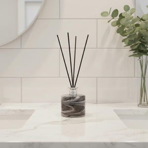 Könnte beinhalten: Ein dekorativer Reed-Diffusor mit schwarzen Stäbchen in einer marmorierten grau-weißen Glasflasche. Der Diffusor steht auf einer weißen Marmor-Arbeitsplatte, mit einem weißen Fliesenhintergrund und einer Vase mit grünem Eukalyptus.
