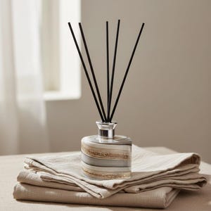May include: A decorative reed diffuser with black reeds and a striped, beige, and gray glass bottle. The diffuser sits atop a stack of folded beige linen. The product is designed for home fragrance.