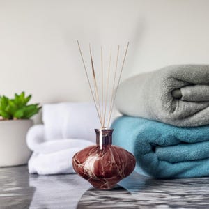 Puede incluir: Una botella difusora de vidrio marrón con remolinos blancos y un juego de cañas de madera. El difusor está sentado sobre una toalla blanca y dos toallas dobladas, una gris y una azul.