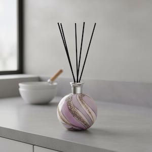 May include: A decorative diffuser with black reeds. The round glass bottle features a swirling pattern of lavender, gold, and white. The diffuser sits on a gray countertop, with a white bowl and window in the background.
