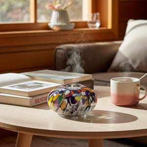 May include: A round, multi-coloured glass candle holder with a speckled pattern of yellow, purple, blue, and white, containing a lit tea light candle that emits a thin wisp of smoke, resting on a light wood surface next to a two-toned mug.