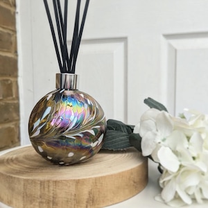 May include: A decorative glass diffuser with black reeds. The round, iridescent glass bottle features a pattern of brown, white, and rainbow colours. The diffuser sits on a wooden slice, with white flowers in the background.