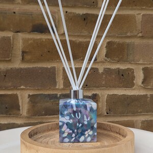 Puede incluir: Un difusor de vidrio azul y blanco con forma cuadrada y un patr&oacute;n decorativo. La botella est&aacute; sentada en una bandeja de madera y tiene palitos difusores blancos.