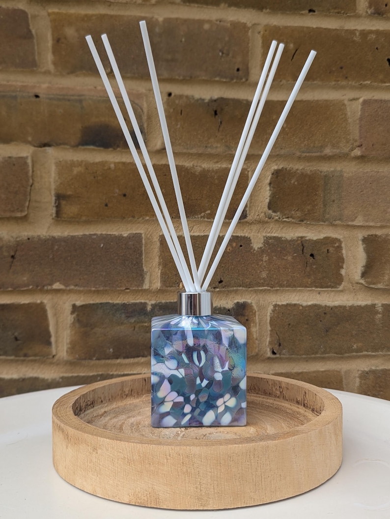 Puede incluir: Un difusor de vidrio azul y blanco con forma cuadrada y un patr&oacute;n decorativo. La botella est&aacute; sentada en una bandeja de madera y tiene palitos difusores blancos.