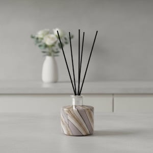 Op de afbeelding: Een decoratieve rietverspreider met zwarte rietjes. De glazen fles heeft een gestreept ontwerp in tinten lavendel, beige en wit. Op de achtergrond staat een kleine witte vaas met witte bloemen.