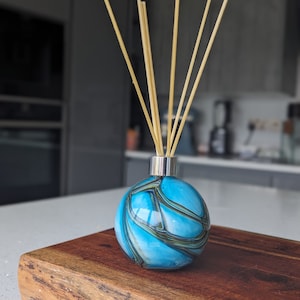 May include: A blue glass diffuser bottle with a silver cap and wooden sticks. The bottle has a swirling pattern of blue, green, and white.