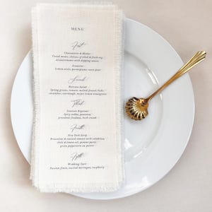 Może przedstawiać: Biały talerz ze złotą łyżką i lnianym menu. Menu zawiera czcionkę w stylu ręcznym z listą dań. Menu znajduje się na białej serwetce z postrzępioną krawędzią.