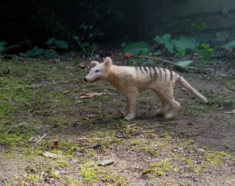 Puede incluir: Un animal de peluche marrón y negro que se asemeja a un tigre de Tasmania, también conocido como tilacino. El animal está de pie sobre una superficie de tierra y hierba.