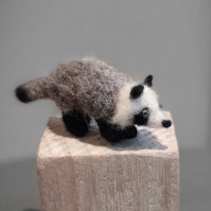 May include: A small, gray and white needle felted badger figurine. The badger is posed in a walking position with its head turned to the side. The figurine is sitting on a wooden block.