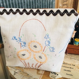 May include: White linen zipped pouch with a brown scalloped trim. The pouch is embroidered with a basket of yellow and orange flowers with blue and green accents.