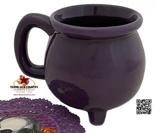 May include: Purple ceramic cauldron mug with a handle and three feet. The mug is on a purple lace doily with a skull and roses design.