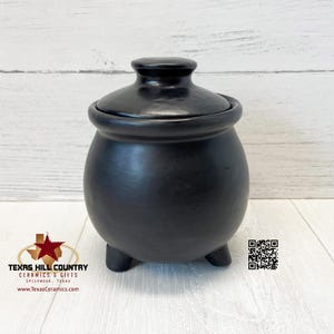 May include: Black ceramic pot with a lid and three legs. The pot is on a white wooden surface.