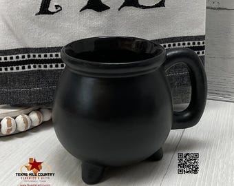 May include: A matte black ceramic mug shaped like a round cauldron with three small feet and a curved handle. The mug is positioned in front of a white fabric with black text and a black and white striped border. A small QR code is visible.