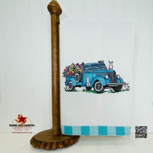May include: A blue vintage pickup truck with a bed full of colorful Easter eggs and bunnies. The truck is embroidered on a white kitchen towel with a blue and white striped border.