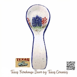 May include: White ceramic spoon rest with a blue and red floral design. The spoon rest has a blue border and is speckled with small black dots. The text "Texas Farmhouse Decor by Texas Ceramics" is at the bottom.
