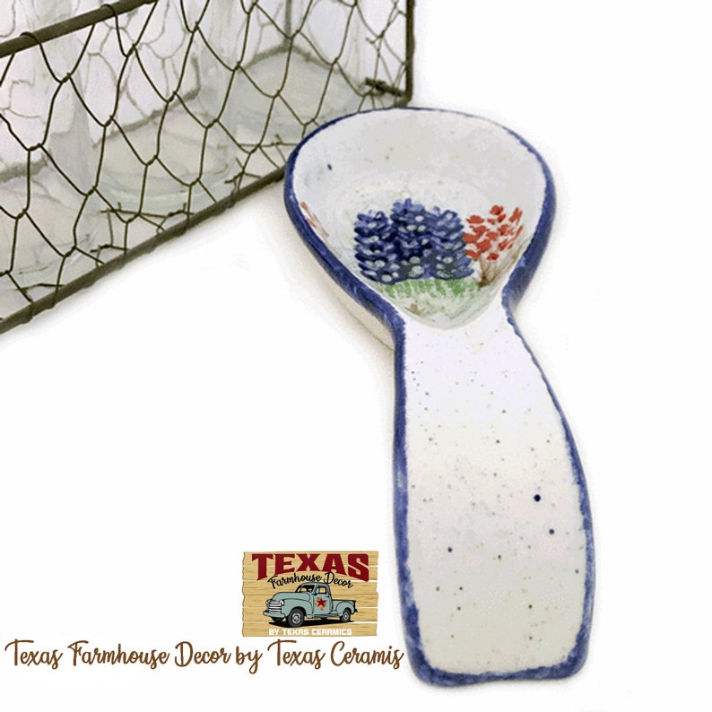 May include: White ceramic spoon rest with a blue rim and a painted floral design. The spoon rest features a bluebonnet design in the center. The text "Texas Farmhouse Decor by Texas Ceramics" is at the bottom.