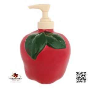 May include: Red ceramic soap dispenser shaped like an apple with a green leaf. The dispenser has a white pump.