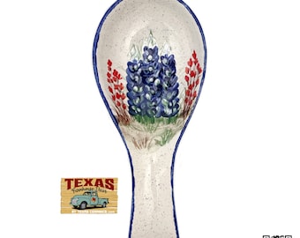 May include: Bluebonnet flower spoon rest with a white background and blue trim. The spoon rest is made of ceramic and has a handle. The spoon rest is decorated with a bluebonnet flower design. The spoon rest is a Texas Farmhouse Decor product.