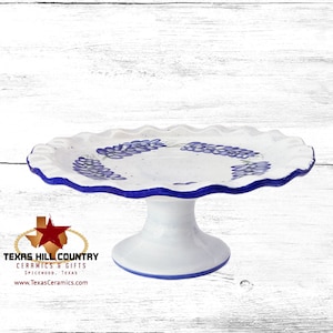 May include: White ceramic cake stand with a blue rim and a blue floral design. The stand has a scalloped edge and a pedestal base.