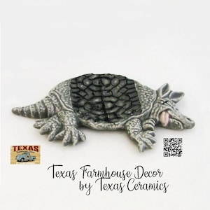 May include: A detailed, gray armadillo figurine with a textured shell and visible claws. The figurine has a pink tongue and is labeled "Texas Farmhouse Decor by Texas Ceramics."