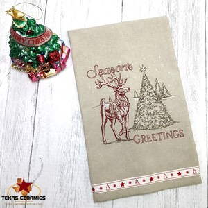 May include: A tan Christmas-themed kitchen towel with a red embroidered design of a reindeer, Christmas tree, and the words "Season's Greetings." The towel has a red and white patterned border with Christmas trees and stars.