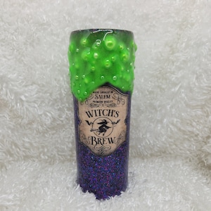 May include: A purple glitter tumbler with a green slime design and a label that reads "Made Locally in Salem, Premium Quality, Witch's Brew".