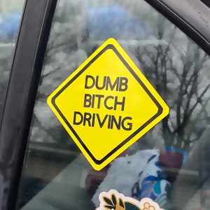 May include: A car window with several stickers. The stickers are yellow, pink, and white. The yellow sticker is a diamond shape with the text "DUMB BITCH DRIVING". The pink sticker is a heart with the text "BAD B*TCH CLUB". The white sticker is a circle with the text "EMBRACE YOUR WORTH".