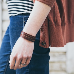 May include: A close-up of a person's wrist wearing a brown leather bracelet with multiple straps. The bracelet is worn over a blue denim jean.
