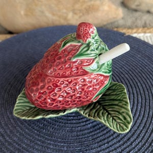 May include: A ceramic strawberry-shaped container with a lid and a small spoon. The container is red with green leaves and a small strawberry on top. The container sits on a green leaf-shaped base.