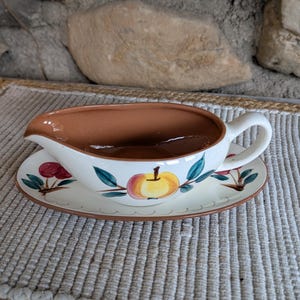 May include: Cream-colored gravy boat and saucer set with a brown interior and handle. The exterior features a hand-painted design of apples and cherries with green leaves. The set is placed on a woven mat.