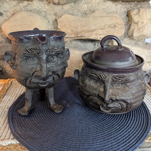 May include: A dark gray ceramic pitcher with three legs and a sculpted face, paired with a matching lidded sugar bowl featuring a similar sculpted face. Both pieces have a glossy finish and detailed facial features including eyes, noses, and mouths.