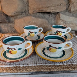 Puede incluir: Cuatro tazas y platillos de cerámica. Las tazas tienen una base blanca con un borde verde y marrón, y un diseño de fruta amarilla. Los platillos tienen un borde amarillo y marrón con un centro verde y blanco. El juego está sobre una estera tejida.