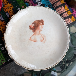 Peut inclure: Une assiette vintage blanc cassé avec un bord festonné et un liseré doré. L'assiette présente un portrait de femme aux cheveux roux et une fleur. L'assiette a un aspect légèrement vieilli et craquelé.