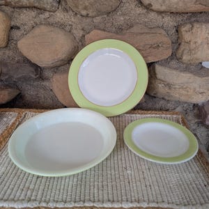 May include: A set of three white ceramic plates with a light green border. The plates vary in size, with a large oval platter, a medium-sized round plate, and a small round plate. The plates are displayed on a woven mat.