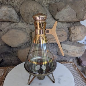 May include: A glass decanter with a conical shape, featuring a metallic gold-tone neck and a matching pointed lid. It has a light beige plastic handle attached to the side and rests on a three-legged metallic base with a gold-tone finish.
