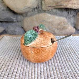 Può includere: Una zuccheriera in ceramica a forma di mela, arancione con foglie verdi e un gambo marrone. Include un cucchiaino d'argento. La zuccheriera misura circa 10 cm di diametro.