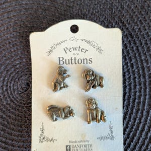 May include: A set of four silver-toned pewter buttons shaped like dogs, attached to a cream-colored display card labeled Pewter Buttons. The card features small illustrations of animals and text reading Handcrafted by DANFORTH PEWTERERS.