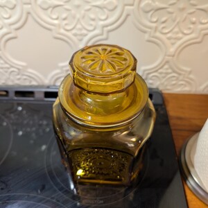 May include: Amber-colored glass jar with a decorative lid. The jar has a geometric shape and a matching lid with a floral design. The jar is sitting on a black surface.