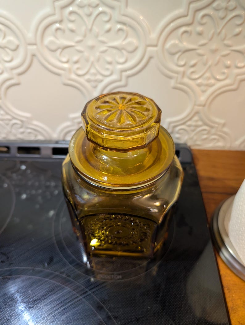 May include: Amber-colored glass jar with a decorative lid. The jar has a geometric shape and a matching lid with a floral design. The jar is sitting on a black surface.