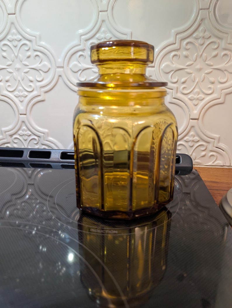 May include: Amber-colored glass jar with a lid. The jar has a hexagonal shape with arched detailing. The lid is a darker shade of amber. The jar is sitting on a black surface.