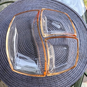 May include: A clear, divided serving dish with a square shape and rounded corners. The edges are outlined in a soft orange color. The dish is divided into three sections, perfect for serving snacks or appetizers.
