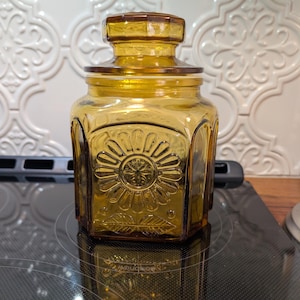 May include: Amber-colored glass jar with a lid. The jar has a hexagonal shape with a floral design on the front. The lid is also amber-colored and has a geometric design. The jar is sitting on a black surface.